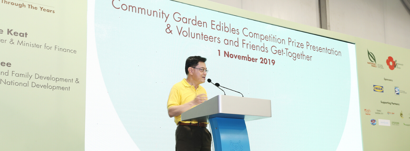 Man at podium with dual mics in front of a screen for "Community Garden Edibles Competition" event. IKEA logo visible.